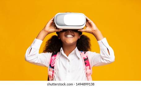 Happy Afro Elementary Student Girl Using VR Headset Experiencing Virtual Reality On Yellow Background. Studio Shot