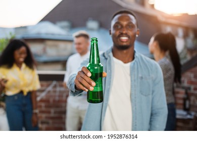 Happy afro man posing on camera with bottle of beer. Blur background of multiracial friends dancing and chatting on open terrace. Party time of young people. - Powered by Shutterstock