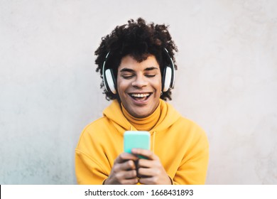 Happy Afro man using mobile smartphone outdoor - Young guy having fun listening music with wireless headphones - Youth millennial generation lifestyle and people addicted technology concept