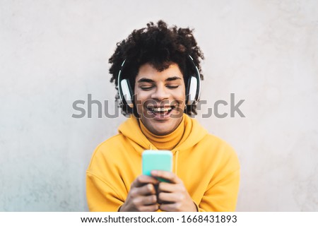 Happy Afro man using mobile smartphone outdoor - Young guy having fun listening music with wireless headphones - Youth millennial generation lifestyle and people addicted technology concept