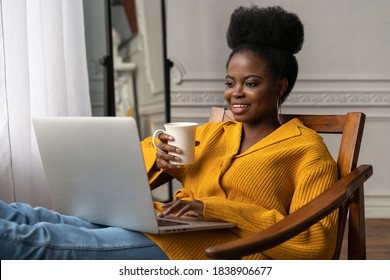 Happy Afro-American Millennial Woman With Afro Hairstyle Wear Yellow Cardigan Resting, Sitting On Chair, Watching Webinar, Working Online On Laptop, Talking In Video Chat, Drinking Tea Or Coffee. 