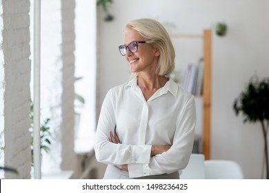 Happy aged grey-haired businesswoman wearing glasses stand with arms crossed look in distance dreaming or visualizing, smiling senior woman worker plan think of success. Business vision concept