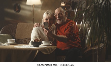 Happy aged husband and wife embracing each other and making video call to friends, via cellphone while sitting on sofa and embracing each other in cozy living room in weekend at home - Powered by Shutterstock