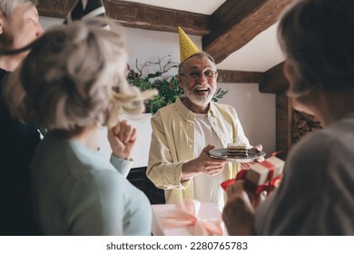 Happy aged man in glasses and party cap standing with birthday cake while friends greeting and giving presents for anniversary celebration - Powered by Shutterstock