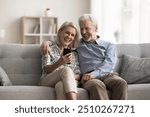 Happy aged wife and husband sit on sofa look at mobile phone screen, laughing, watching funny videos in social media, spend carefree pastime on internet using gadget. Olde gen and modern tech usage