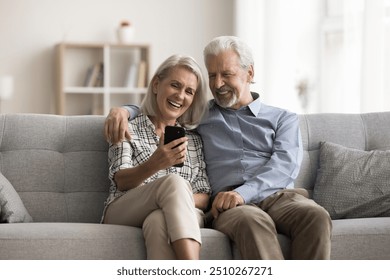 Happy aged wife and husband sit on sofa look at mobile phone screen, laughing, watching funny videos in social media, spend carefree pastime on internet using gadget. Olde gen and modern tech usage - Powered by Shutterstock