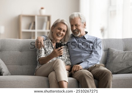 Happy aged wife and husband sit on sofa look at mobile phone screen, laughing, watching funny videos in social media, spend carefree pastime on internet using gadget. Olde gen and modern tech usage