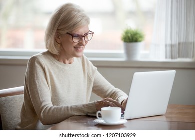 Happy aged woman in glasses working at laptop drinking tea, smiling senior female using computer browsing or surfing internet, reading news online, excited elderly lady texting message at pc at home