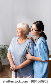 Happy Aged Woman With Walking Stick And Young Asian Nurse Smiling And Looking Away In Hospital