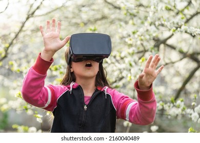 Happy Amazed Little Girl Wearing Virtual Reality Goggles Watching Movies Or Playing Video Games. Cheerful Surprised Child Looking In VR Glasses And Gesturing