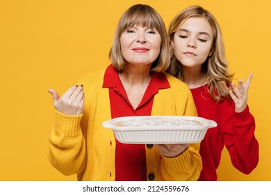 Happy Amazed Woman 50s In Red Shirt Have Fun With Teenager Girl 12-13 Years Old. Grandmother Granddaughter Hold Meal Dish Casserole Sniff Isolated On Plain Yellow Background. Family Lifestyle Concept