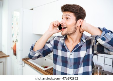 Happy amazed young man in checkered shirt talking on cell phone on the kitchen - Powered by Shutterstock