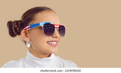 Happy American Child On Beige Copyspace Studio Background. Cute Little Girl Wearing Sunglasses With US Flag Design Looking At Blank Text Space Background On Right Side. Kids Fashion, July 4th Concept