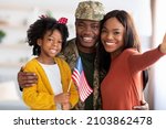 Happy American Soldier Taking Selfie With Family After Returning Home From Army, Black Man In Military Uniform Posing At Camera With Wife And Little Daughter Holding USA Flag, Closeup Shot, POV