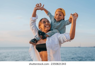 Happy American  woman in white shirt plays with little son sitting piggyback looks at camera happily against ocean on summertime vacations. Leisure, family moments, sincere people emotions. Rest. - Powered by Shutterstock