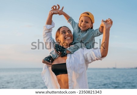 Happy American  woman in white shirt plays with little son sitting piggyback looks at camera happily against ocean on summertime vacations. Leisure, family moments, sincere people emotions. Rest.