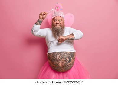 Happy Amused Bearded European Man Has Fun On Carnival Or Costume Party Dances With Raised Arms Holds Magic Wand Wears Fairy Outfit Has Fat Tattooed Belly. People Holiday Celebration Concept.