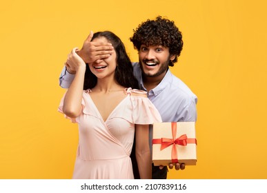 Happy anniversary. Loving indian man covering his girlfriend's eyes, greeting her with wrapped gift box, posing over yellow studio background, celebrating special day