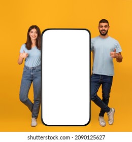 Happy Arab Couple Standing By Huge Cellphone With Blank Screen And Showing Thumb Ups, Recommending Newest Mobile App Or Website Ad, Mockup, Yellow Studio Background