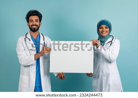 Happy arab doctors man and woman in uniform and hijab showing white blank placard for advertisement or text, muslim medical workers showing copy space, standing on blue studio background, mockup