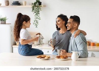 Happy Arab Family Enjoying Breakfast At Cozy Kitchen, Little Girl Daughter Sitting On Table, Drinking Milk And Having Conversation With Hugging Smiling Father And Mother Drinking Morning Coffee