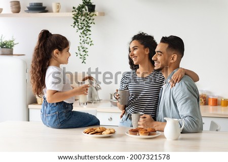 Happy arab family enjoying breakfast at cozy kitchen, little girl daughter sitting on table, drinking milk and having conversation with hugging smiling father and mother drinking morning coffee
