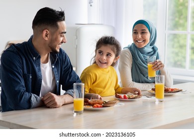 Happy Arab Family Of Three Eating Tasty Lunch Together At Home, Cheerful Islamic Parents And Little Daughter Sitting At Table In Kitchen, Enjoying Healthy Delicious Food And Drinking Orange Juice