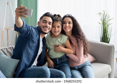 Happy Arab Family Of Three Taking Selfie With Smartphone At Home, Cheerful Middle Eastern Parents And Cute Little Daughter Posing For Photo While Relaxing Together On Couch In Living Room