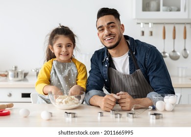 Happy Arab Father And Little Daughter Baking Cookies In Kitchen Together, Adorable Girl Kneading Dough While Cooking With Dad At Home, Middle-eastern Family Wearing Aprons And Smiling At Camera