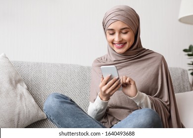 Happy arab female in hijab scrolling social networks or watching photo on smartphone at home, sitting on comfortable sofa, free space
