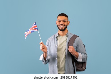 Happy Arab Guy With Backpack Showing Flag Of Great Britain Over Blue Studio Background, Copy Space. Positive Middle-eastern Male Student Studying English At School