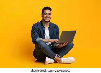 Happy arab guy in casual freelancer using modern laptop, working online, yellow studio background. Cheerful middle eastern young man independent contractor sitting on floor with notebook, copy space - Shutterstock ID 2107608107