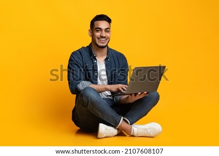 Happy arab guy in casual freelancer using modern laptop, working online, yellow studio background. Cheerful middle eastern young man independent contractor sitting on floor with notebook, copy space