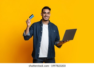 Happy Arab Guy With Laptop And Credit Card On Yellow Studio Background, Positive Middle-eastern Young Man Holding Bank Card And Brand New Computer, Shopping Or Banking Online, Copy Space