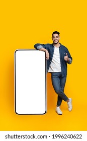 Happy arab guy leaning on huge smartphone with empty screen and showing thumb up, mockup, yellow studio background, full length shot, copy space, mobile application, advertisement concept - Shutterstock ID 1996240622