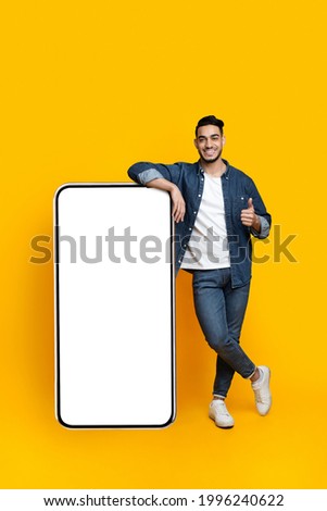 Happy arab guy leaning on huge smartphone with empty screen and showing thumb up, mockup, yellow studio background, full length shot, copy space, mobile application, advertisement concept