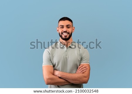 Happy arab guy smiling to camera, crossing hands and posing over blue background, studio shot. Excited self-confident middle eastern man expressing positive emotions