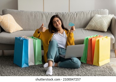Happy Arab Lady Shopping Online From Laptop, Using Credit Card, Sitting On Floor With Paper Bags And Showing Thumb Up. Excited Middle Eastern Woman Buying Goods From Home