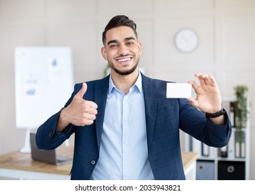 Happy Arab Male Entrepreneur Holding Empty Business Card, Showing Thumb Up Gesture At Company Office, Mockup For Design. Smiling CEO Manager Demonstrating Blank Advertising Card