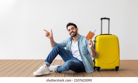 Happy Arab Male Tourist Holding Plane Tickets And Passport, Pointing At Free Space And Smiling At Camera, Sitting Near Suitcase Over Light Wall, Panorama With Copy Space