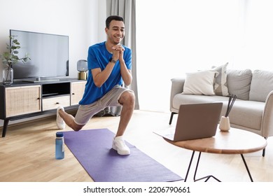 Happy Arab Man Doing Forward Lunge Exercises While Watching Online Workout Tutorial Via Laptop, Training At Home. Domestic Male Fitness Concept