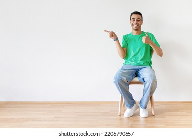 Happy Arab Man Gesturing Thumb Up And Pointing Finger Aside At Free Space, Sitting On Chair Over Light Wall Background. I Like This Offer And Promo Concept