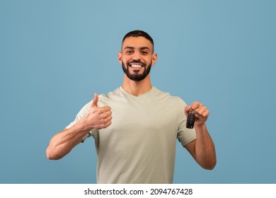 Happy arab man holding car key and showing thumb up, smiling at camera while posing over blue background in studio. Excited guy enjoying buying new car - Powered by Shutterstock