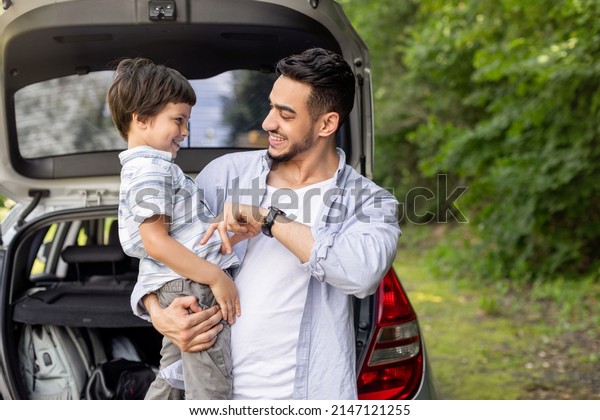 Happy arab man holding son in arms and playing with\
him near car with open trunk outdoor in summer. Family travel,\
vacation together and auto journey at free time, picnic outside in\
park, copy space