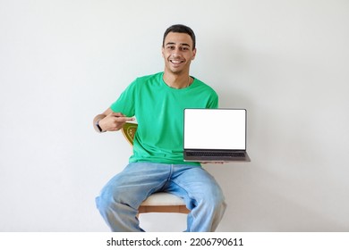 Happy Arab Man Pointing At Laptop Pc With Empty Screen, Sitting On Chair Against Light Studio Wall, Mockup For Website. Young Guy Presenting Portable Computer With Ad Template