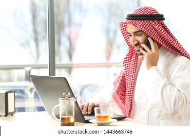 Happy Arab Man Using A Laptop And Talking On Smart Phone Sitting In A Coffee Shop