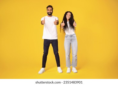 Happy arab millennial man and woman in casual showing thumb ups and smiling at camera, posing full length, showing like gestures with both hands standing over yellow background, studio shot - Shutterstock ID 2351483901