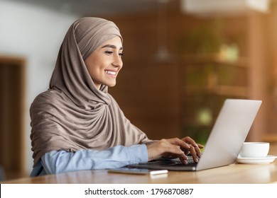 Happy arab woman freelancer chatting with clients on laptop, sitting at cafe, empty space