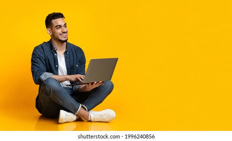 Happy Arab Young Man Sitting On Floor, Using New Modern Laptop, Typing On Keyboard And Smiling Over Yellow Studio Background, Looking At Copy Space For Text Or Advertisement, Panorama