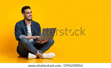 Happy arab young man sitting on floor, using new modern laptop, typing on keyboard and smiling over yellow studio background, looking at copy space for text or advertisement, panorama